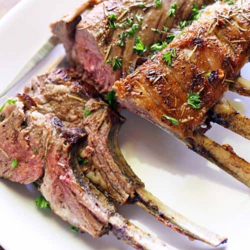 Roasted rack of lamb is topped with chopped parsley and served on a white plate.