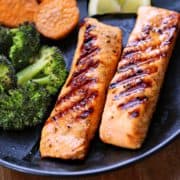 Grilled salmon is served on a dark plate with broccoli and sweet potatoes.