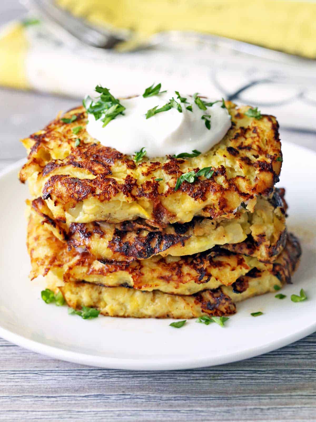 Yellow squash fritters are stacked on a white plate and topped with sour cream.