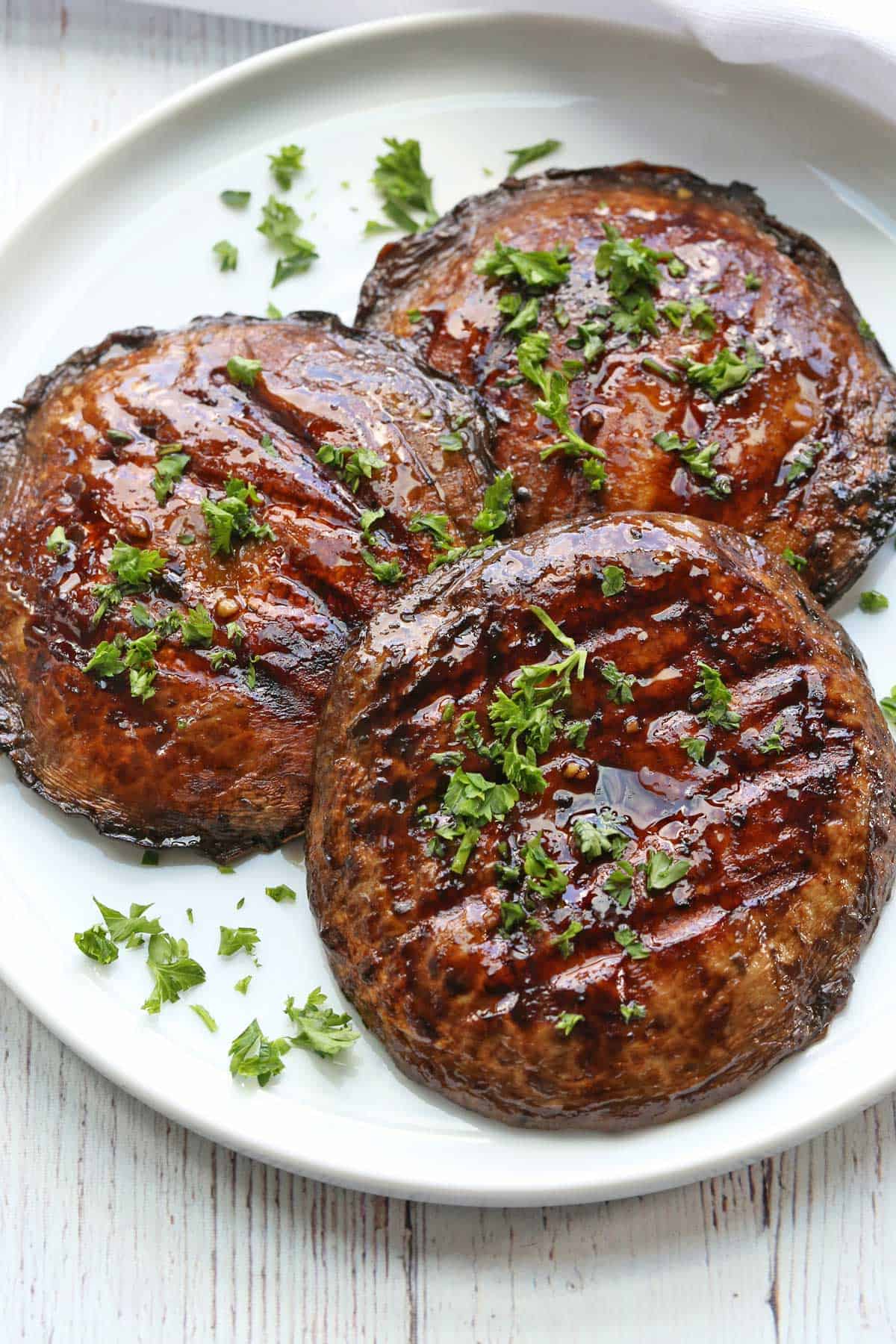Three grilled portobello mushrooms are served on a white plate, topped with parsley. 