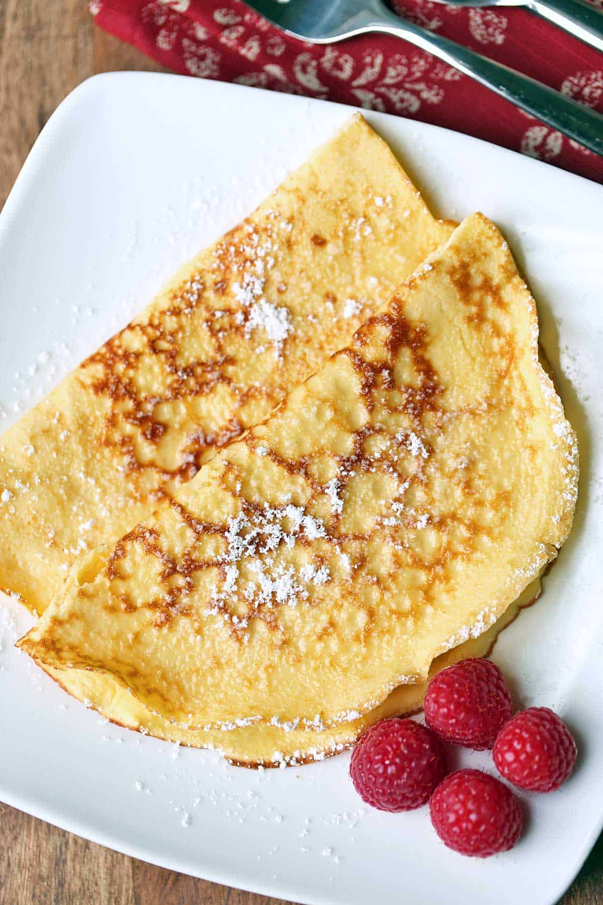 Two keto crepes are served on a white plate garnished with raspberries. 