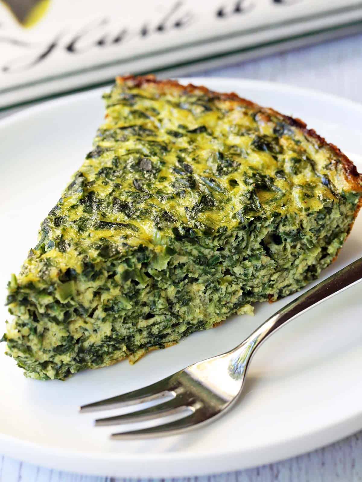 A slice of a crustless spinach quiche is served on a white plate with a fork.