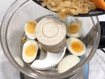 Adding sauteed onions to food processor. There are hard-boiled eggs in the food processor.