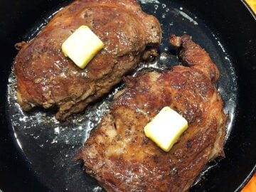 Two ribeye steaks in a cast-iron skillet, topped with butter.