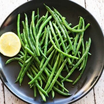 Haricot verts are served on a black plate with a slice of lemon.