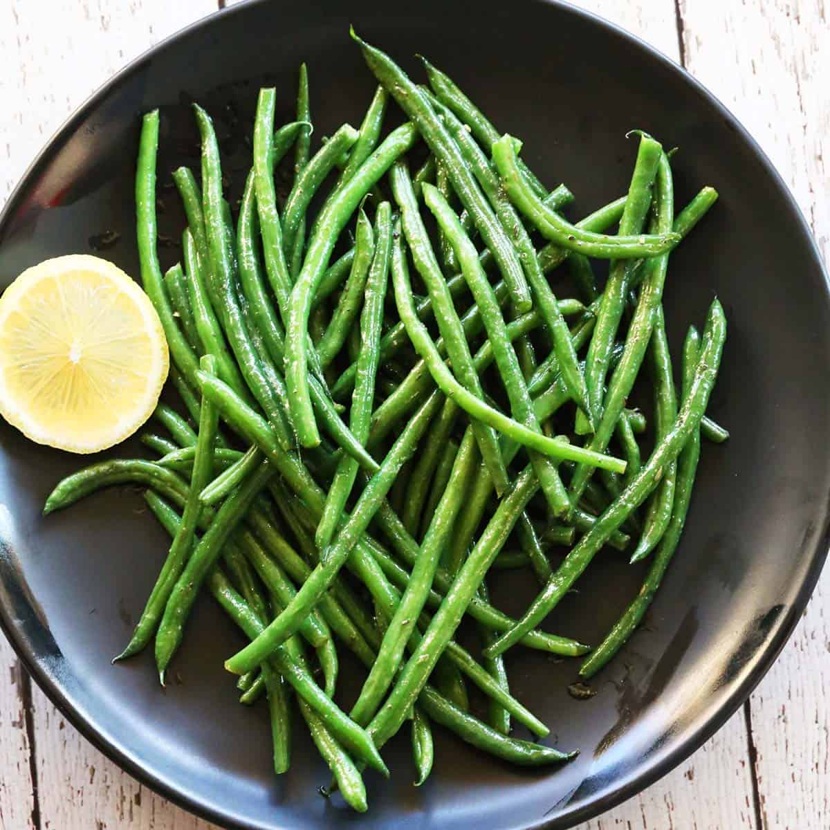 Haricot verts are served on a black plate with a slice of lemon.