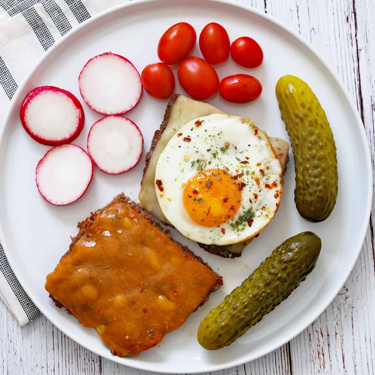 Two baked hamburgers are served on a white plate with veggies and pickles.