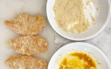 Three coated chicken tenders on a cutting board next to bowl of egg and almond flour.