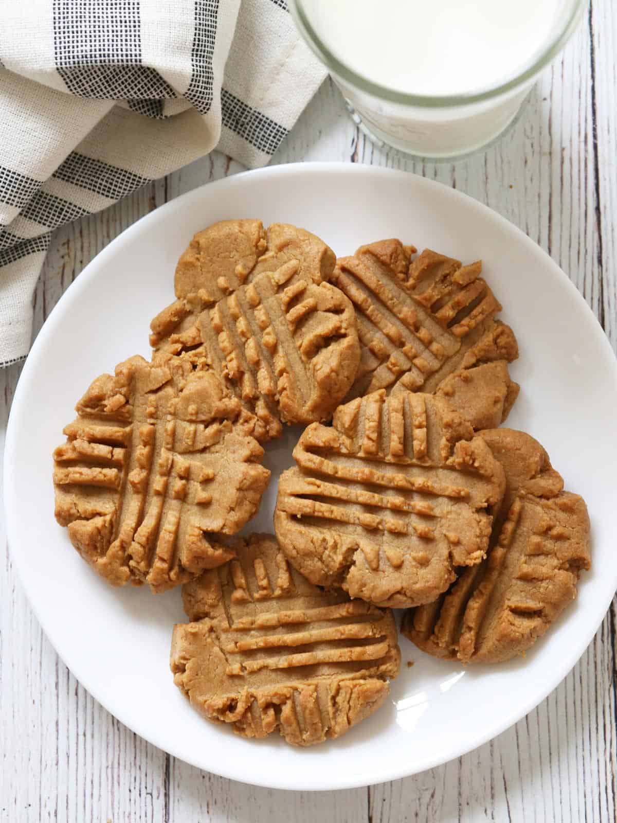 Keto peanut butter cookies are served on a white plate with a napkin and a glass of milk.