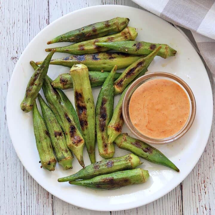 Grilled okra is served on a white plate with a dipping sauce.