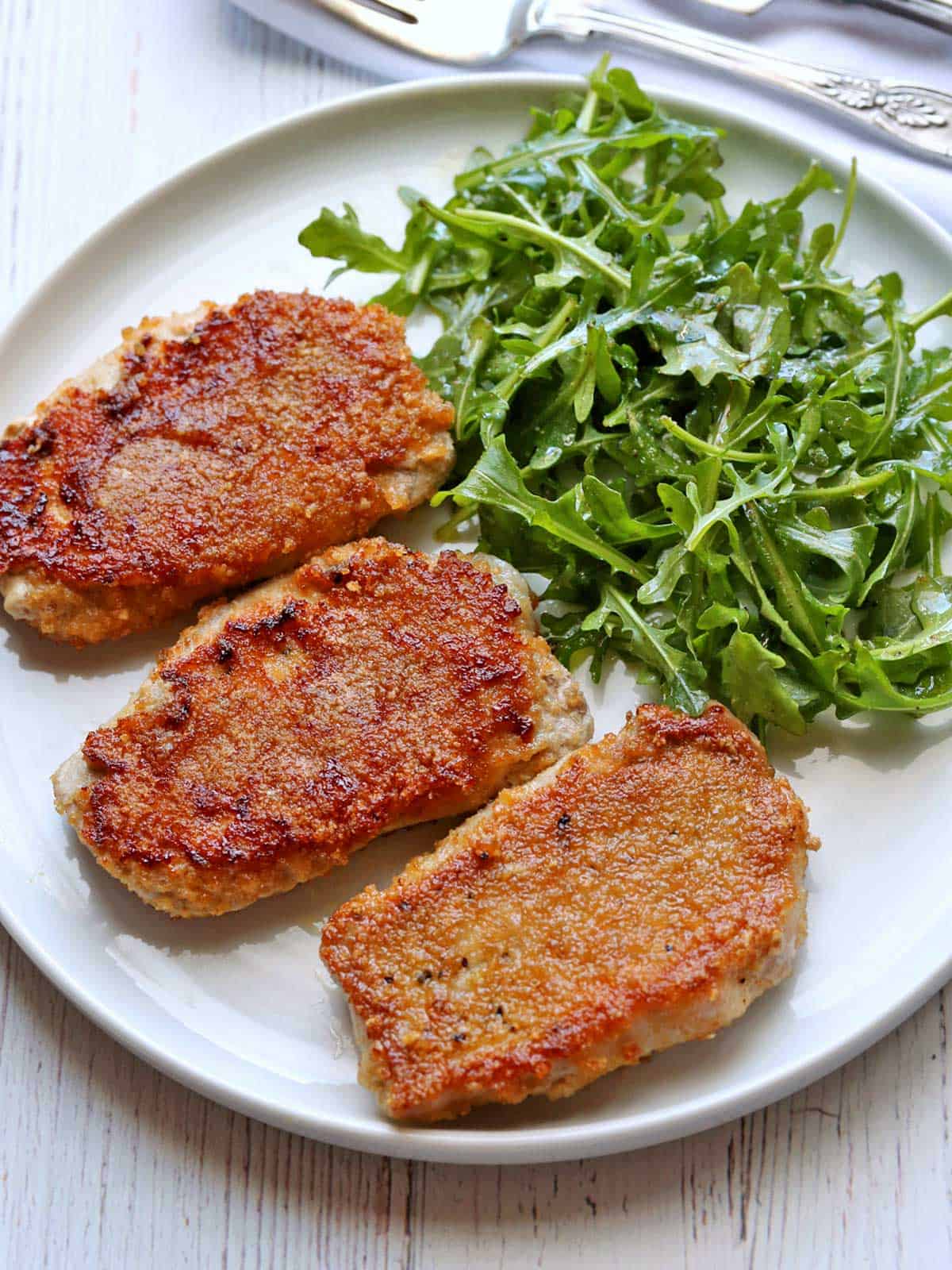 Three keto fried pork chops are served with arugula salad.