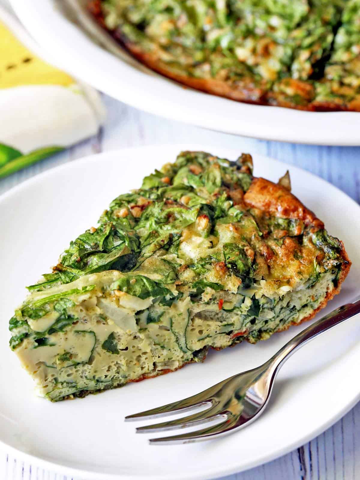 A slice of spinach frittata is served on a plate. A pie plate with the entire frittata is shown in the background.