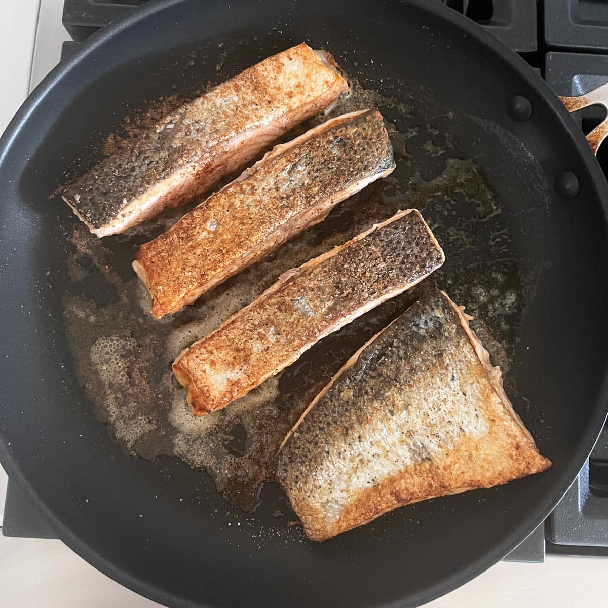 Four salmon fillets are being pan-fried in a skillet. One of them is wider and thinner than the others.
