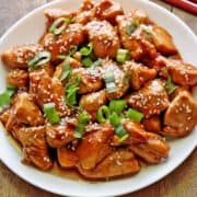 Keto sesame chicken is served on a white plate, topped with sesame seeds and scallions.