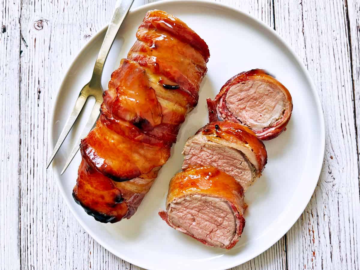 Bacon wrapped pork tenderloin is served on a white plate.