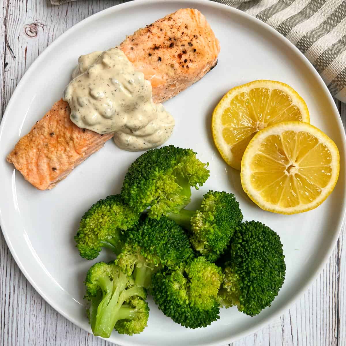 Poached salmon is topped with tartar sauce and served with steamed broccoli and lemon slices.