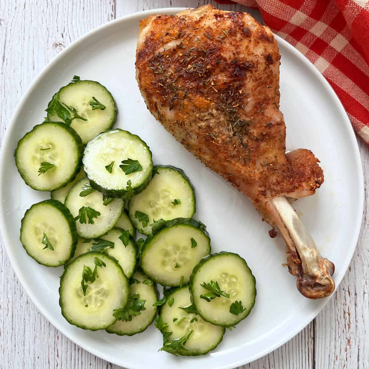 A roasted turkey leg is served on a white plate with cucumber salad.