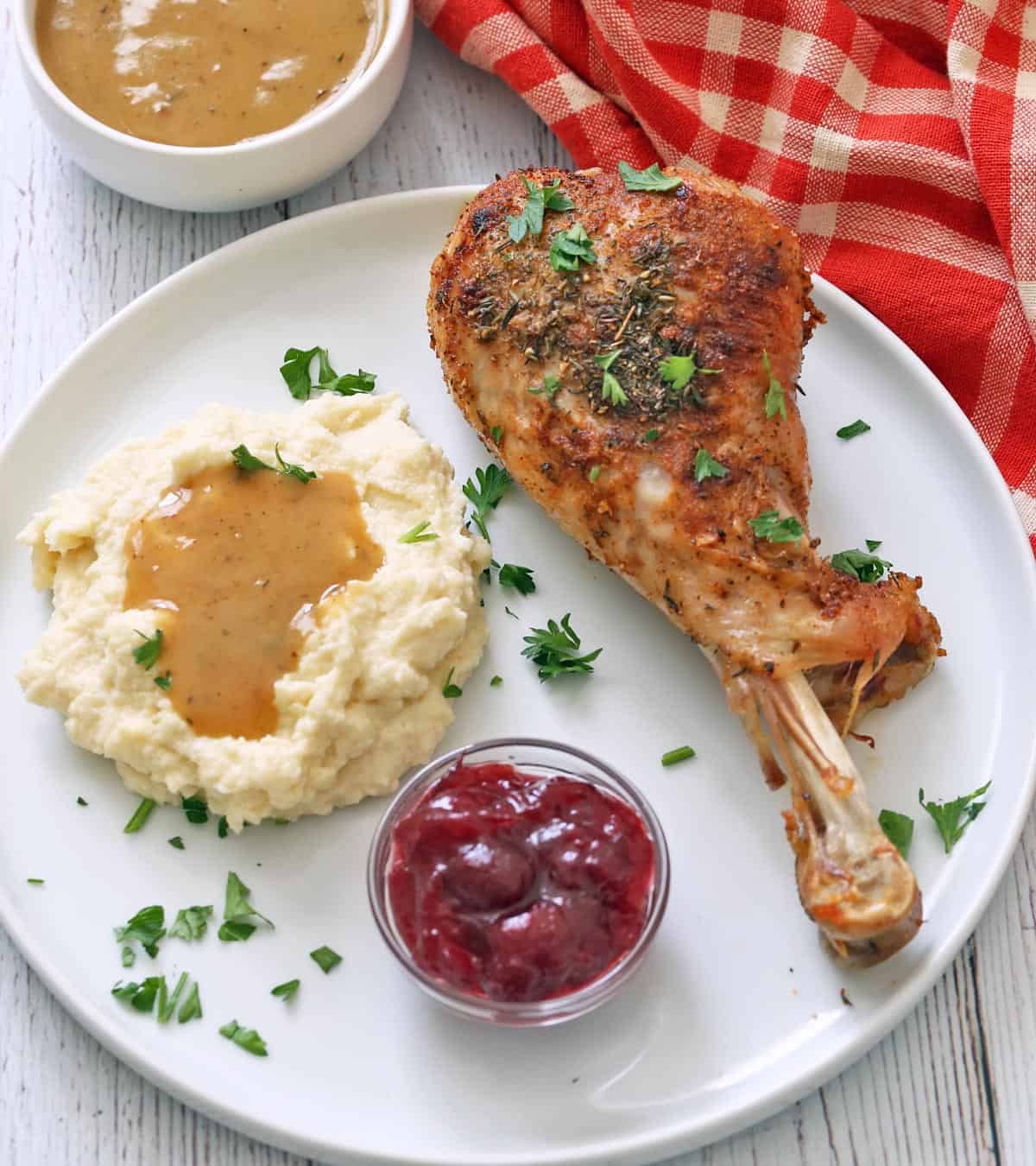 A roasted turkey leg is served on a plate with mashed cauliflower, gravy, and cranberry sauce.