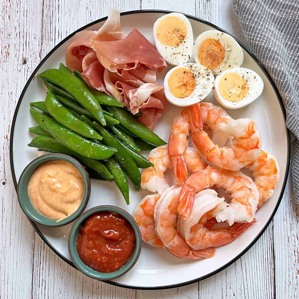 A plate with shrimp, cocktail sauce, sriracha mayo, prosciutto, hard-boiled eggs, and sugar snap peas.