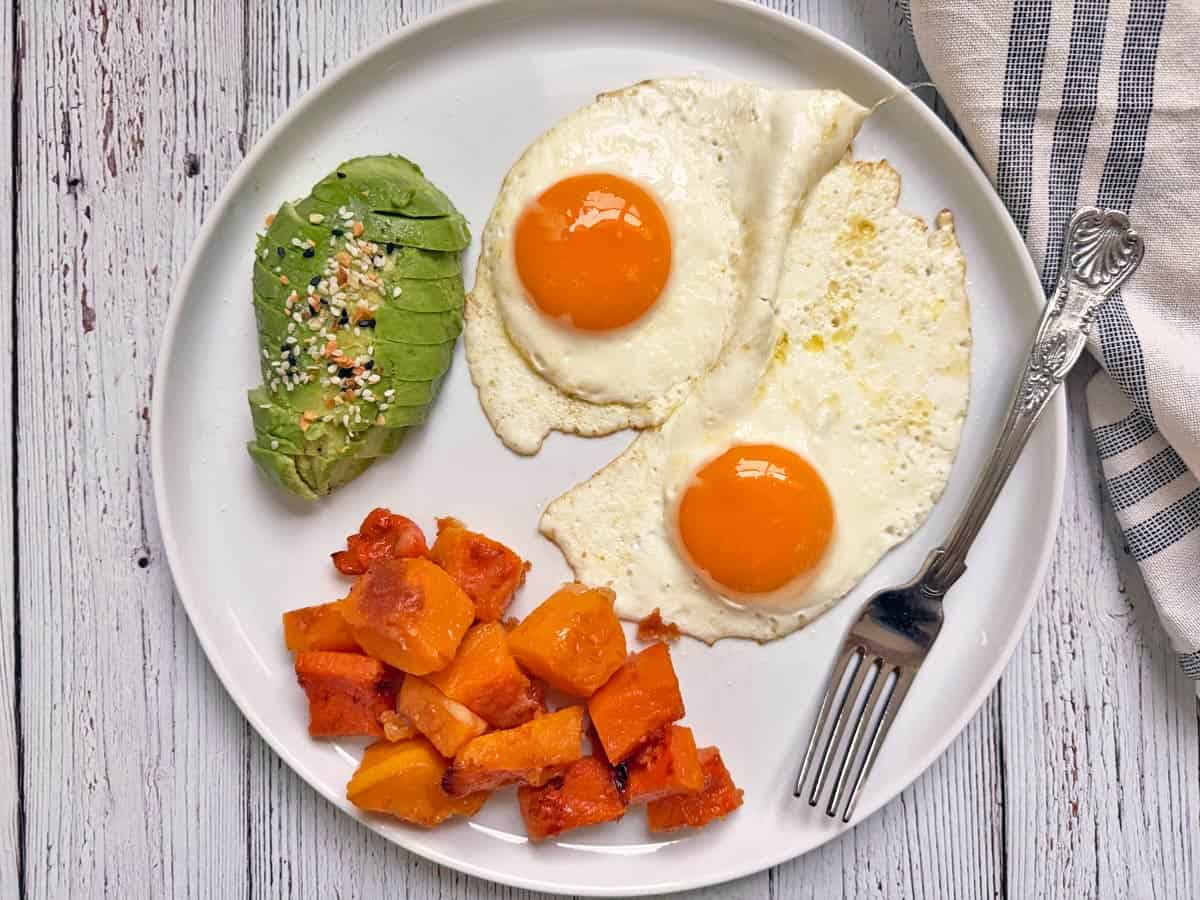A white plate with fried eggs, avocado, and butternut squash.